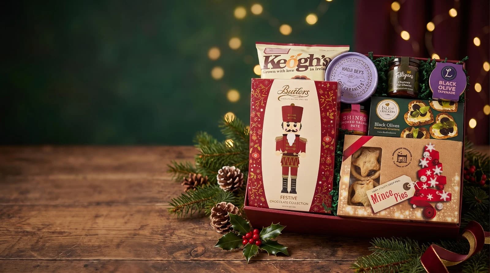 A Christmas hamper on a wooden table, holding a Butlers festive chocolate collection, Keogh’s crisps, Hadji Bey’s caramels, a Filligan’s chutney, Shines smoked salmon pâté, Isle of Crackers and a box of mince pies, with pine, holly and pinecones around it