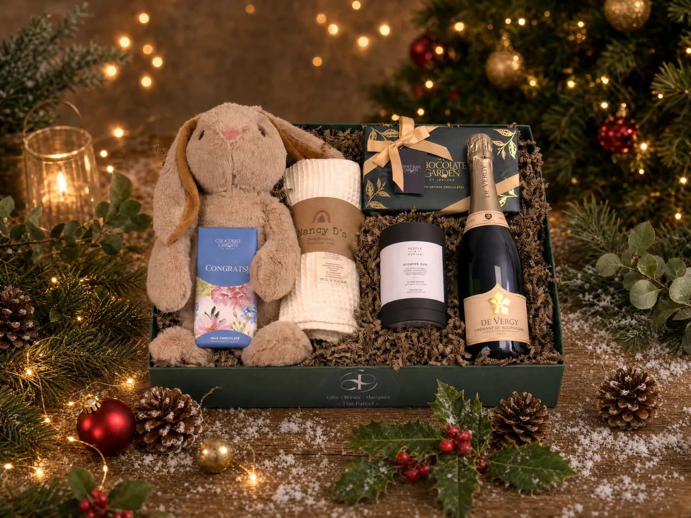 The New Baby Celebration hamper dressed for Christmas, a plush bunny and a rolled blanket beside a congratulations chocolate bar and a bottle of sparkling wine