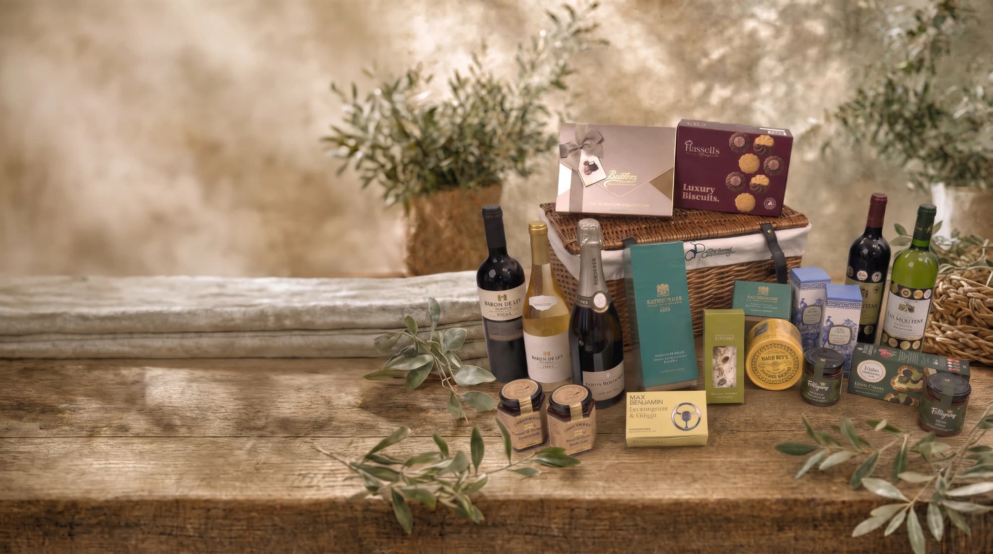 A wicker hamper on a wooden table with red, white and sparkling wines, Butlers chocolates, luxury biscuits, preserves and candles laid out in front of it